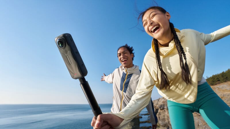 Two people smiling and posing with their arms outstretched while taking a photo with a 360-degree camera on a selfie stick, standing on a rocky coastline with sky and ocean in the background.