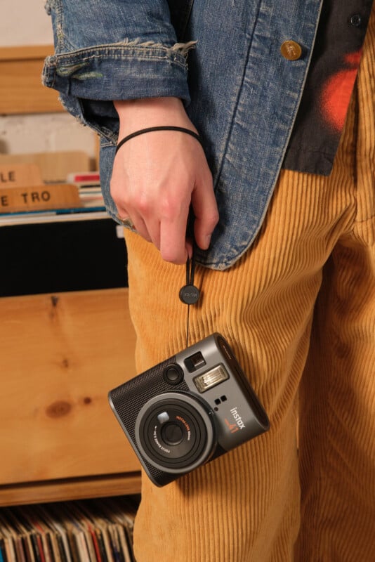 Person in a denim jacket and corduroy pants holding a black Fujifilm Instax camera. They are standing by a wooden cabinet with records in the background.