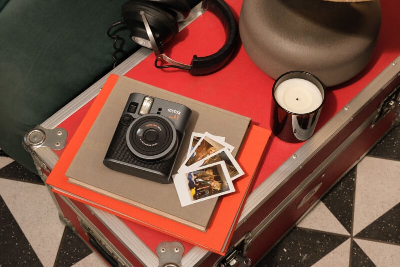 Instant camera with photos on notebooks, placed on a red table. Nearby are headphones, a lit candle, and a red and silver storage case. The floor has a black and white geometric pattern.