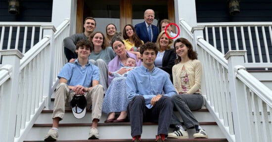 A multigenerational family of twelve, including a baby, poses and smiles together on the front steps of a house. A man in a suit stands at the top; a woman behind him is circled in red.
