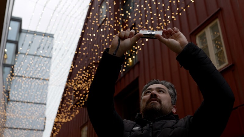 A person with a beard holds a camera above their head, capturing a scene of twinkling lights overhead. They stand between tall buildings with string lights creating a festive atmosphere. The person wears a dark jacket, looking upwards.