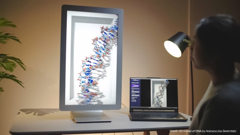 A person looks at a large computer screen displaying a 3D DNA model, with a laptop showing the same model beside it on a desk. A lamp and a plant are also visible in the softly lit room.