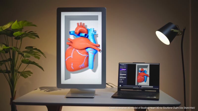 A 3D animated model of a human heart is displayed on a large vertical screen, with a laptop beside it showing the same image. A lamp and a plant are on the white desk in a well-lit room.