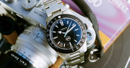 A stainless steel wristwatch with a black face and luminous markings is placed on a vintage camera. The watch has two sub-dials and a branded logo. The background includes a colorful, blurred book cover.