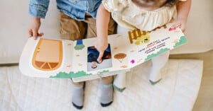 Two young children sit side by side on a quilt, looking at an open illustrated book. The page features colorful shapes and drawings. One child is pointing to a picture on the page. The children are wearing casual clothing and socks.