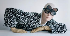 Person in a black and white patterned coat poses against a white background. They wear round, black goggles and tan gloves, with one hand placed on a white surface.