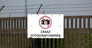 A weathered sign on a metal fence displays a crossed-out camera icon and the text "Zakaz Fotografowania," indicating no photography allowed. The fence is topped with barbed wire, and greenery is visible in the background.