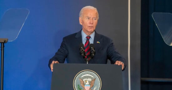 A man in a navy suit speaks at a podium with microphones. The podium displays the United States presidential seal. A blue background and teleprompters are visible.
