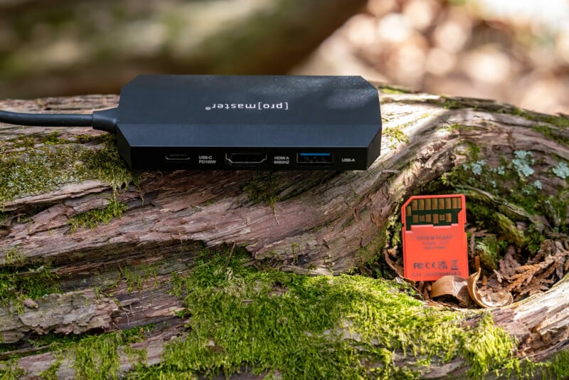 A black multi-port hub with USB and HDMI slots rests on a moss-covered piece of wood. Next to it is an orange SD card partially inserted into a crevice in the wood. Background is blurred, showing natural textures.