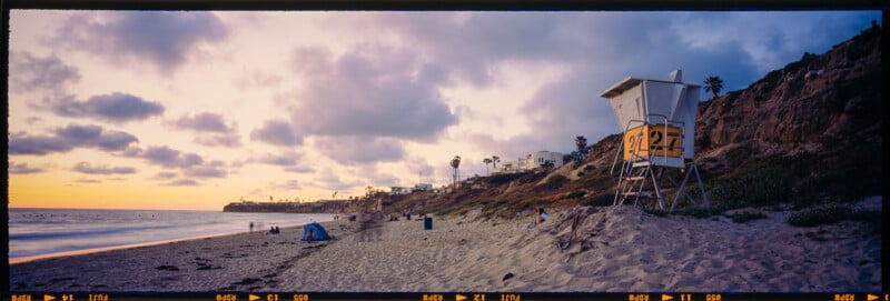 Beach scene at sunset with a lifeguard tower marked "27" on the right. The sky is cloudy and the coastline is lined with cliffs and buildings. There's a tent on the sand in the distance, creating a peaceful atmosphere.