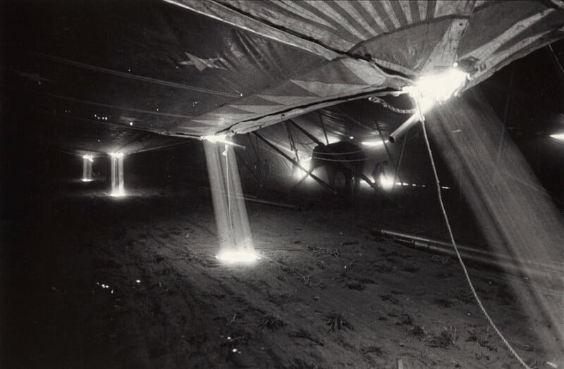 Sunbeams stream through holes in the roof of a large circus tent, illuminating dust in the air and casting light on the ground below. The scene is mostly dark, with bright, dramatic shafts of light.