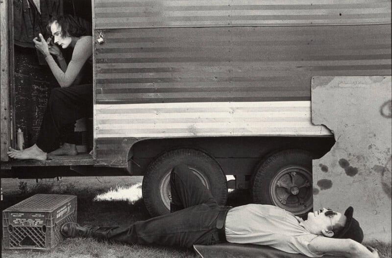 A person sits in the doorway of a striped trailer, applying makeup, while another person lies on the grass below, resting with a cap over their eyes. A crate is placed nearby outside the trailer.