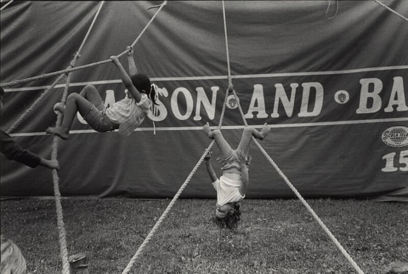 Two children play on rope structures in front of a large circus tent. One child hangs upside down by their knees, while the other climbs the ropes. The ground below is grassy. Part of a circus sign is visible in the background.