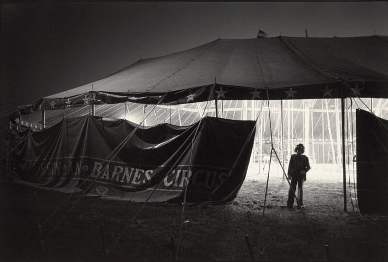 A person stands in front of a large, illuminated circus tent at night, with a dark banner partially reading "BARNES CIRCUS" draped across the entrance and light shining from within.