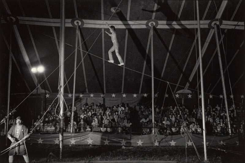 A tightrope walker balances high above a seated circus audience under a large tent, while a person on the ground watches closely. The tent is decorated with stars, and spotlights illuminate the scene.