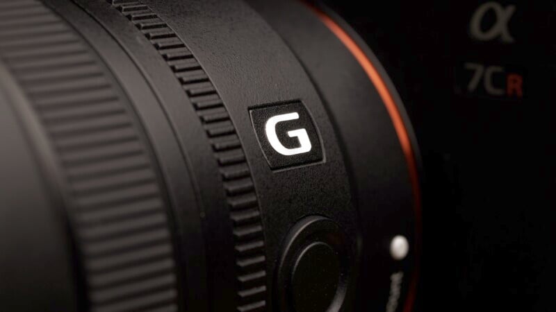 Close-up of a camera lens with a prominent "G" logo, featuring a textured focus ring and part of the camera body visible, highlighted by soft lighting.