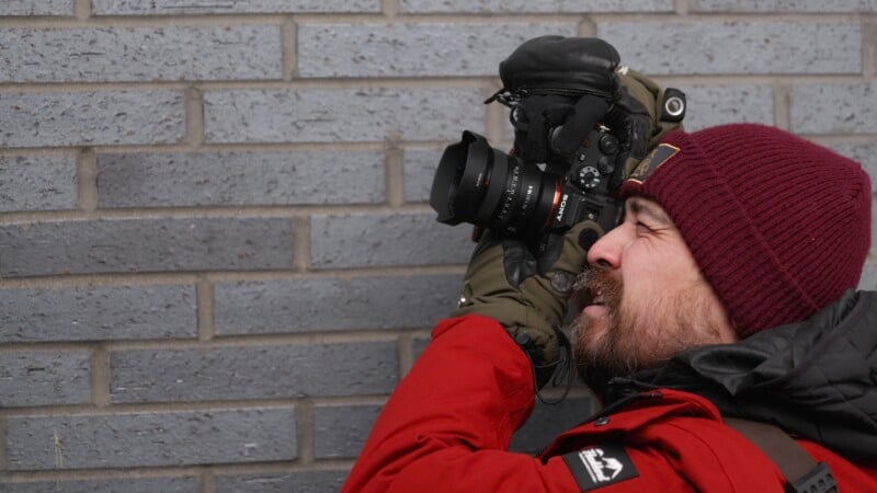 A person in a red jacket and maroon beanie is holding a camera up to their eye, aiming at a gray brick wall. The person is wearing green gloves and appears focused on taking a photo.
