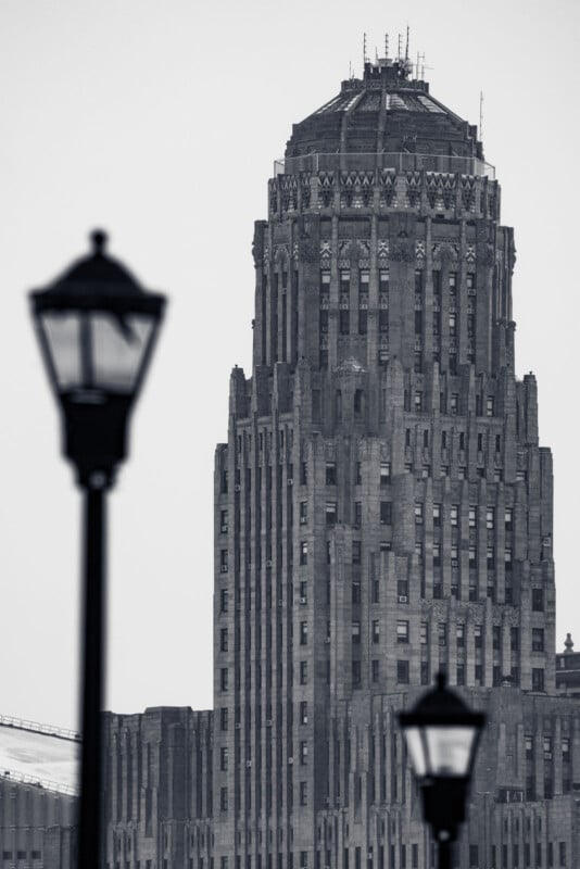 Black and white photo of a tall, intricate Art Deco skyscraper. In the foreground, two blurred street lamps frame the building. The sky is overcast, enhancing the dramatic, vintage feel of the architectural scene.
