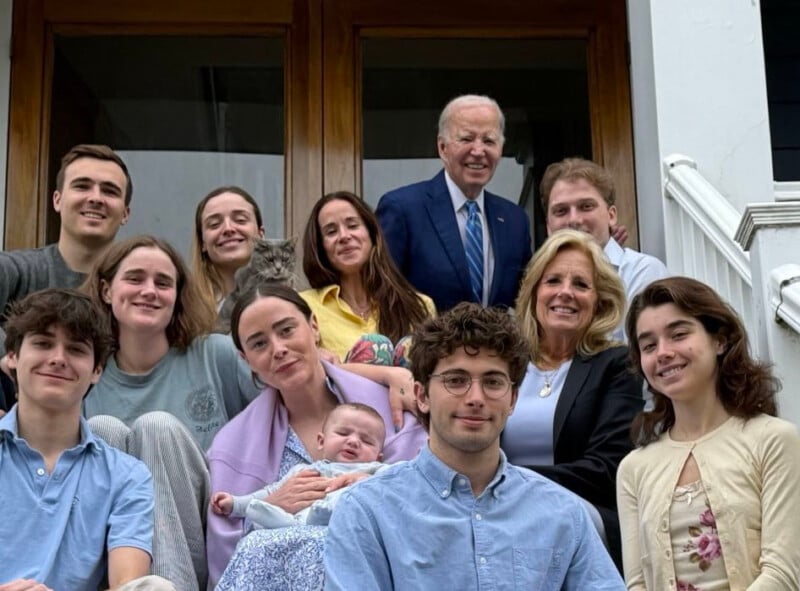 A group of people, including an older man in a suit and woman in a blazer, pose together on outdoor steps in front of a wooden door. Some are seated, some standing, and one person holds a baby. All are smiling at the camera.