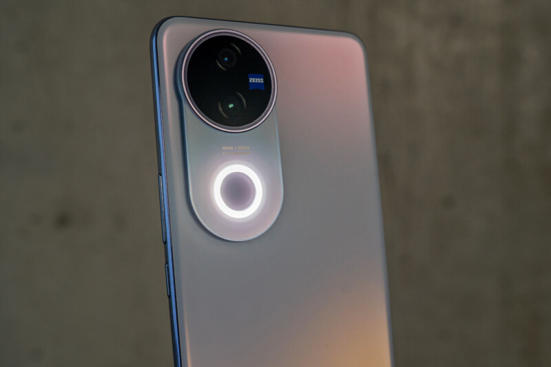 Close-up of a smartphone's rear camera module, featuring a large circular design with two lenses and a small text that says "Vivo ZEISS." The phone has a sleek, metallic finish, and the background is a blurred gray surface.