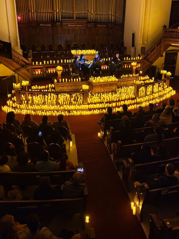 A dimly lit concert in a church features musicians on stage surrounded by numerous glowing candles. The audience sits in pews, watching the performance. Ornate wooden architecture and organ pipes are visible in the background.