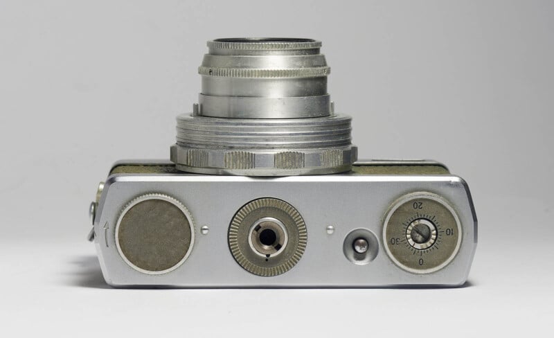 A close-up view of the top of a vintage silver camera with a protruding lens, dials, and knobs against a plain light background.