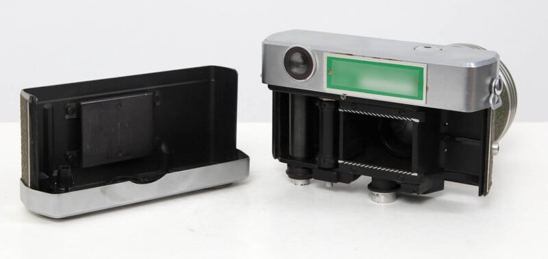 A vintage film camera with its back cover removed, showing the inner film compartment and winding mechanisms, resting on a white surface.