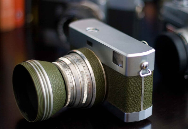 A vintage camera with a green textured body and metallic lens sits on a dark surface, surrounded by blurred cameras in the background.