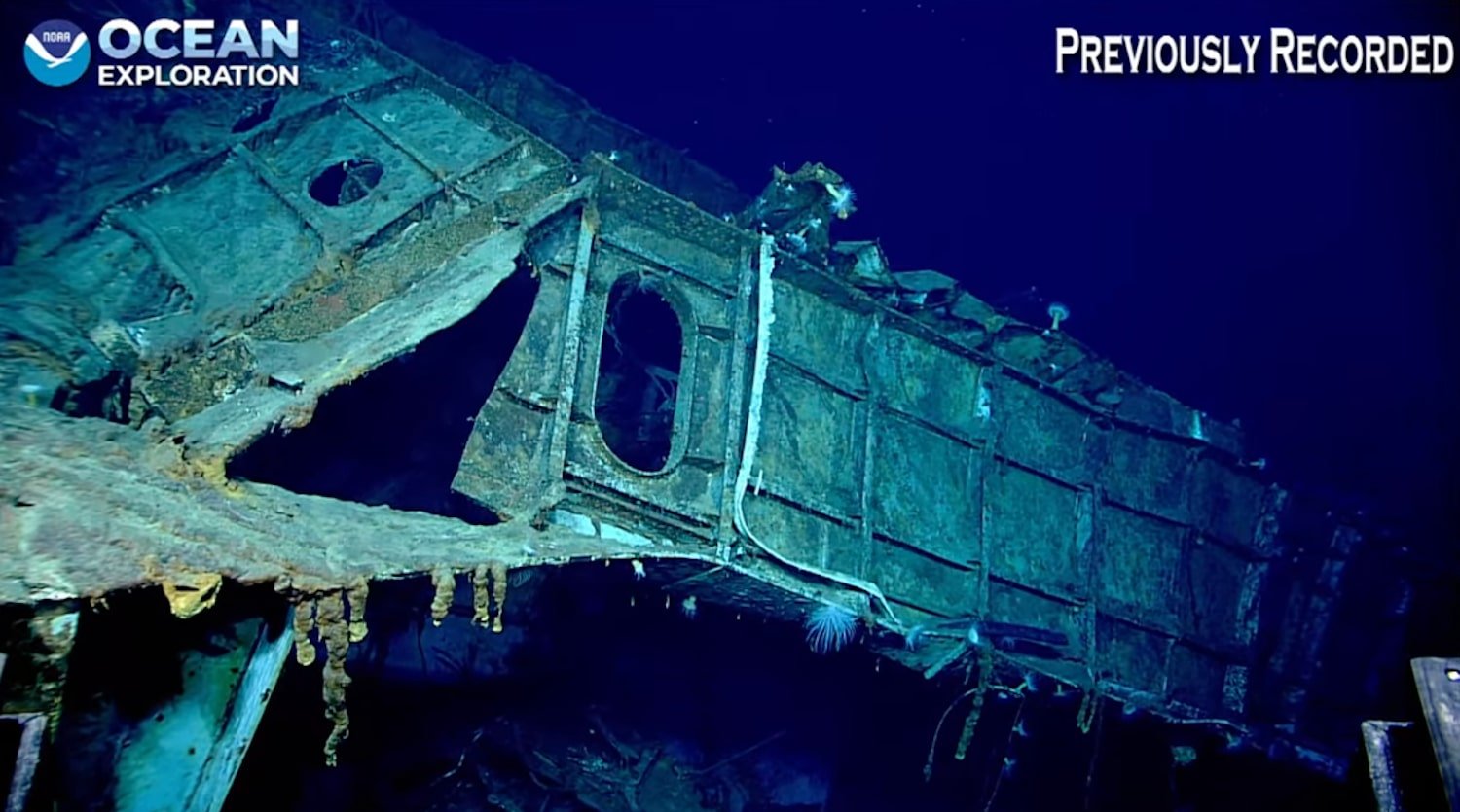A rusted, broken section of a shipwreck rests on the seafloor in deep blue water. The image includes the NOAA Ocean Exploration logo and the words "Previously Recorded" in the top corners.
