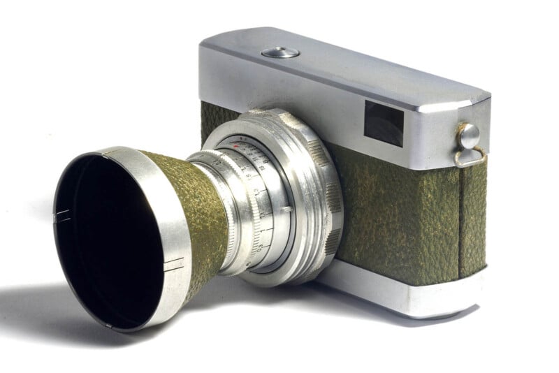 A vintage film camera with a silver metal body and olive green textured grip, featuring a prominent lens with a large hood, is shown on a white background.