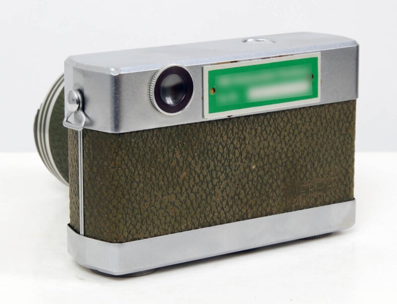 A vintage camera with a textured green and silver body, a prominent lens on the left, and a green rectangular label on the top right front. The camera is displayed against a plain, light background.