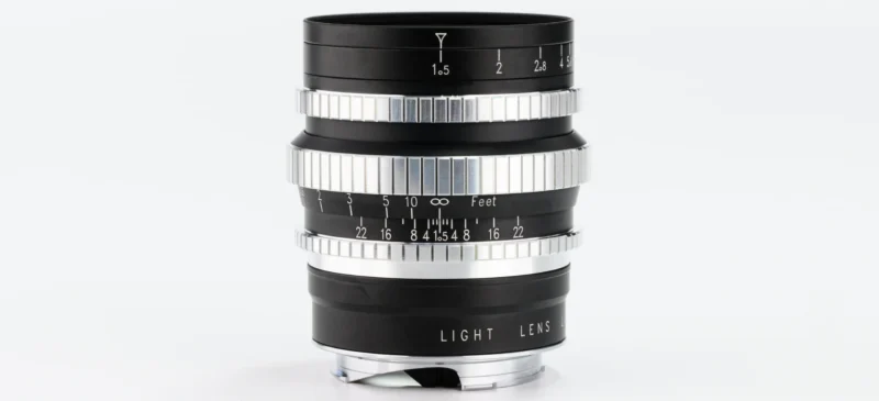 A black and silver camera lens with distance and aperture markings is displayed upright against a plain white background. The lens features knurled focus and aperture rings for adjustment.