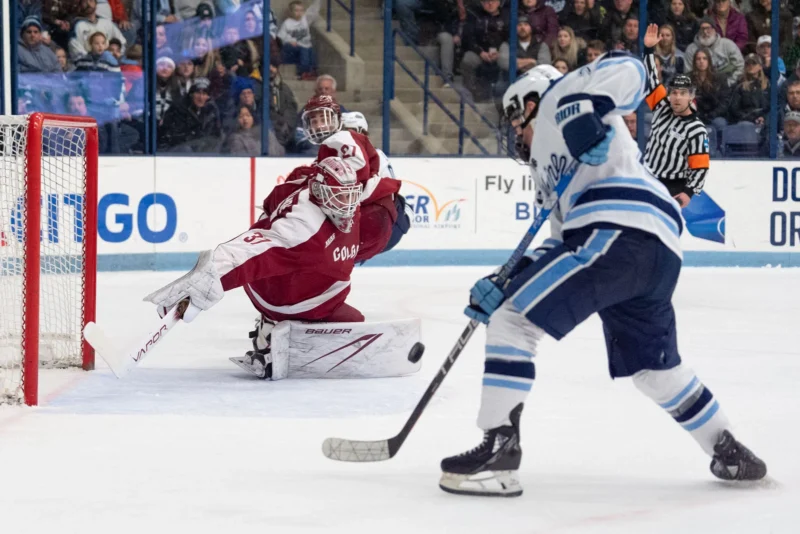 A hockey goalie in red gear dives to block a shot as a player in blue and white prepares to shoot the puck toward the net; fans watch from the stands in the background.